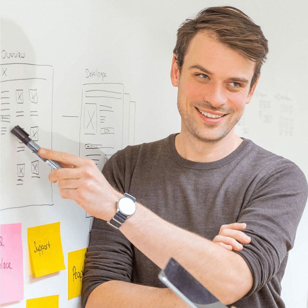 A man is explaining something in front of a whiteboard covered with sketches and notes related to portal development. He is holding a marker and smiling as he points to one of the drawings. The image conveys a creative and collaborative work atmosphere in the field of portal development. Portal Development with SUNZINET